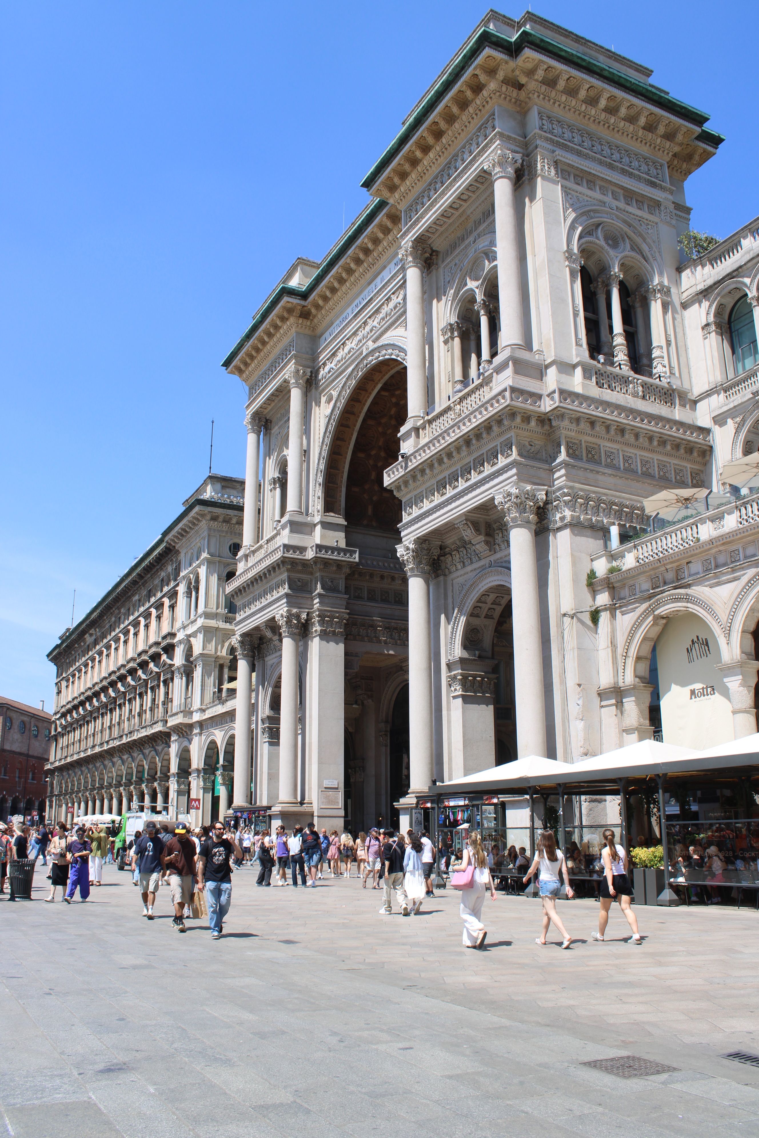 Galleria Vittorio Emanuele II entrance bustling with people on a sunny day in Milan.