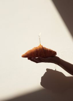 A hand holding a croissant with a lit candle casting dramatic shadows on a plain wall.