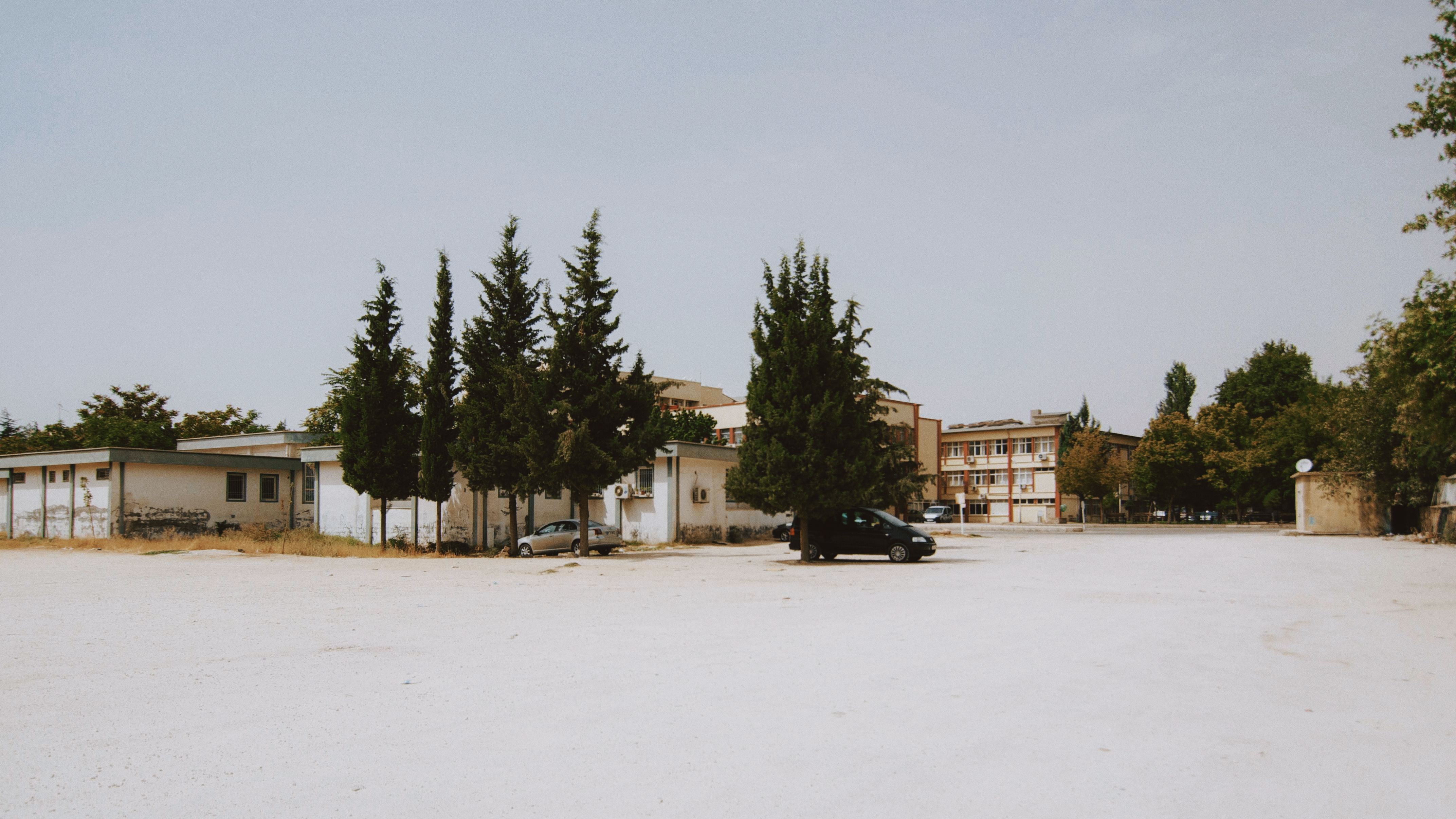 Wide shot of an empty urban lot with buildings and tall trees under a clear sky.