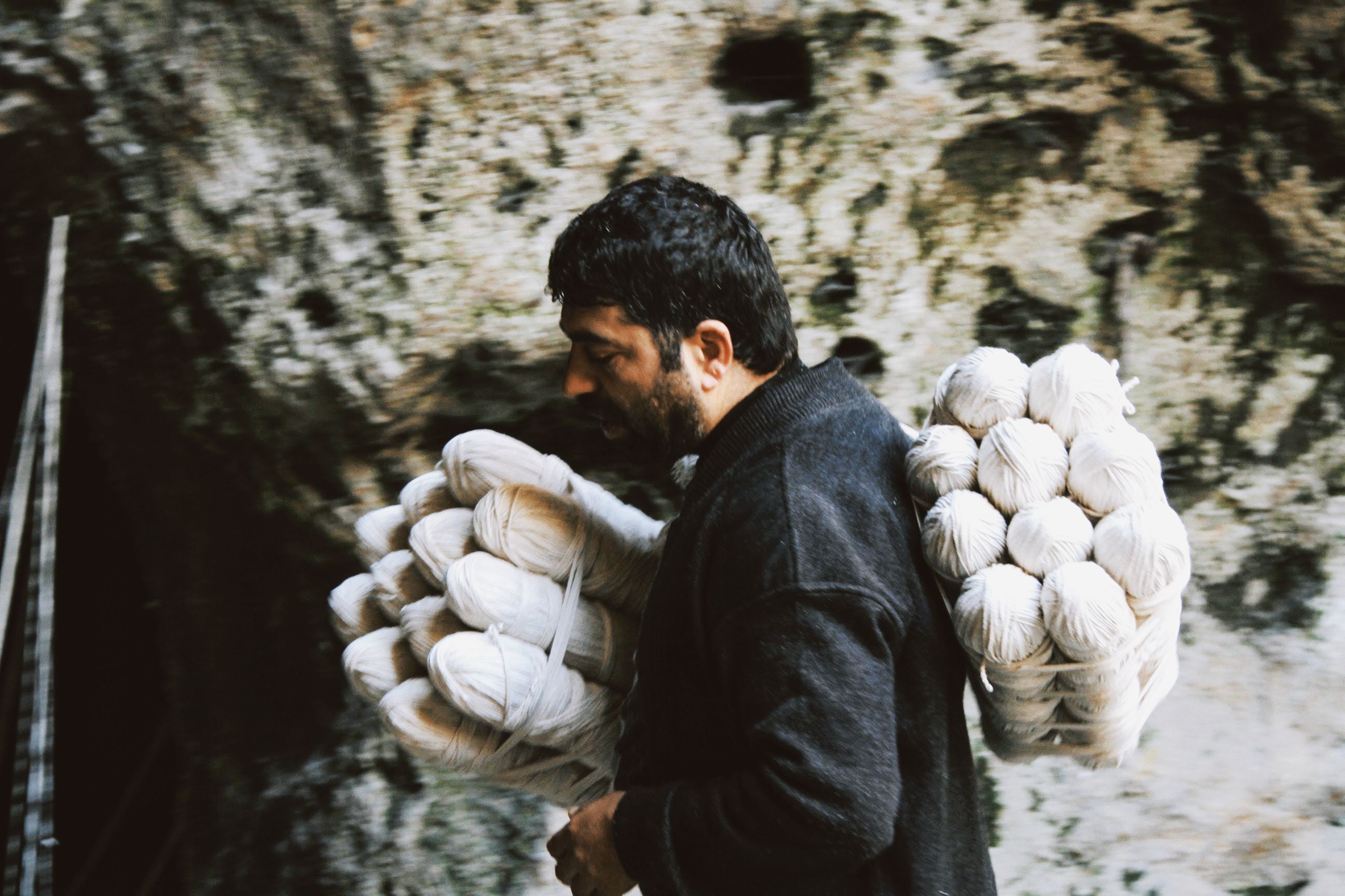 Bearded man carrying bundles of yarn against a natural rock wall backdrop.