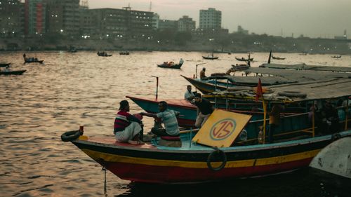 Free Men interacting on colorful boats by the riverside as evening falls, city skyline in the background. Stock Photo