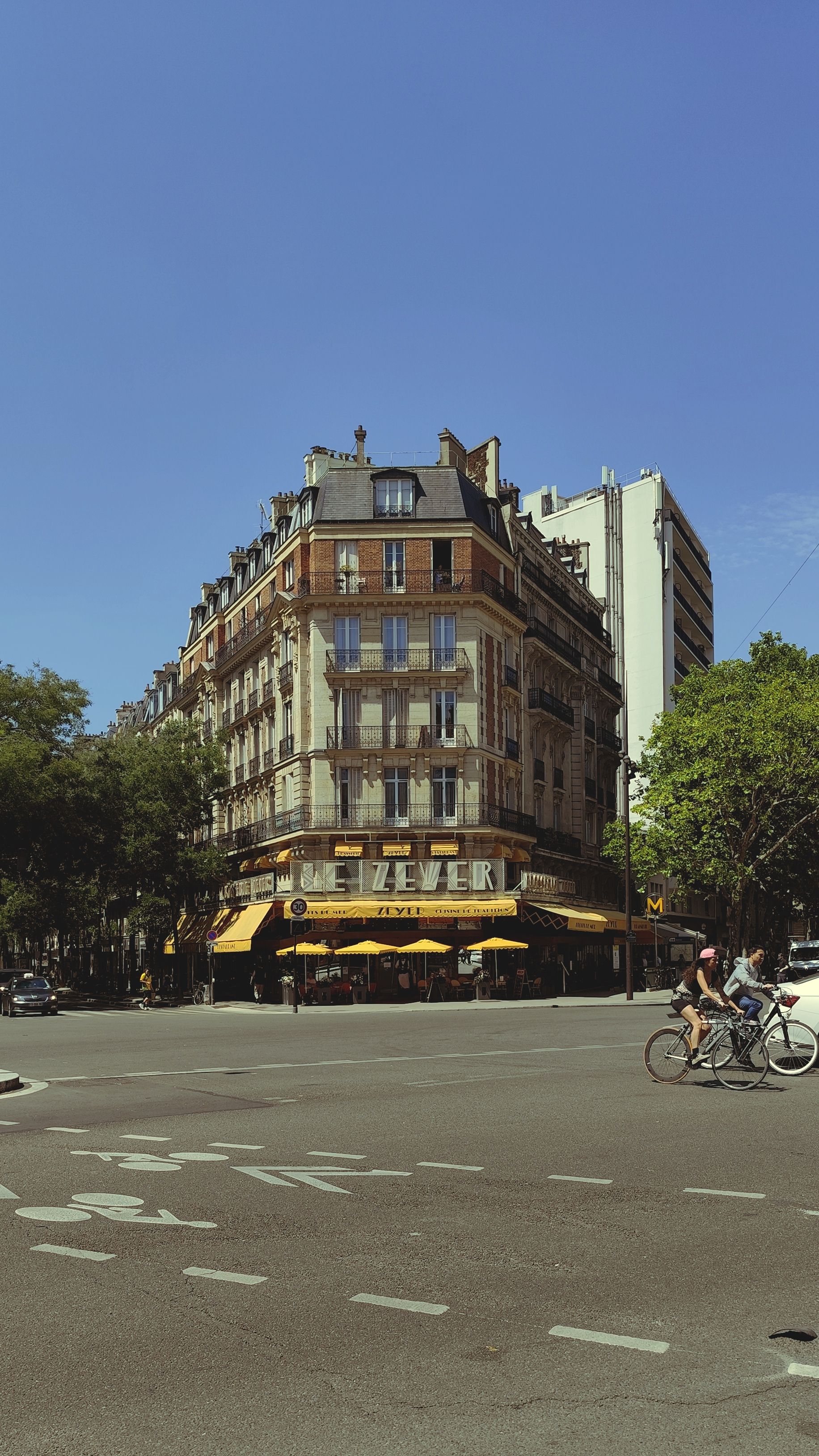Charming Parisian corner cafe with cyclists passing by under a clear blue sky.