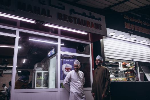 Two men in traditional attire enjoy the vibrant nightlife at a Muscat restaurant.