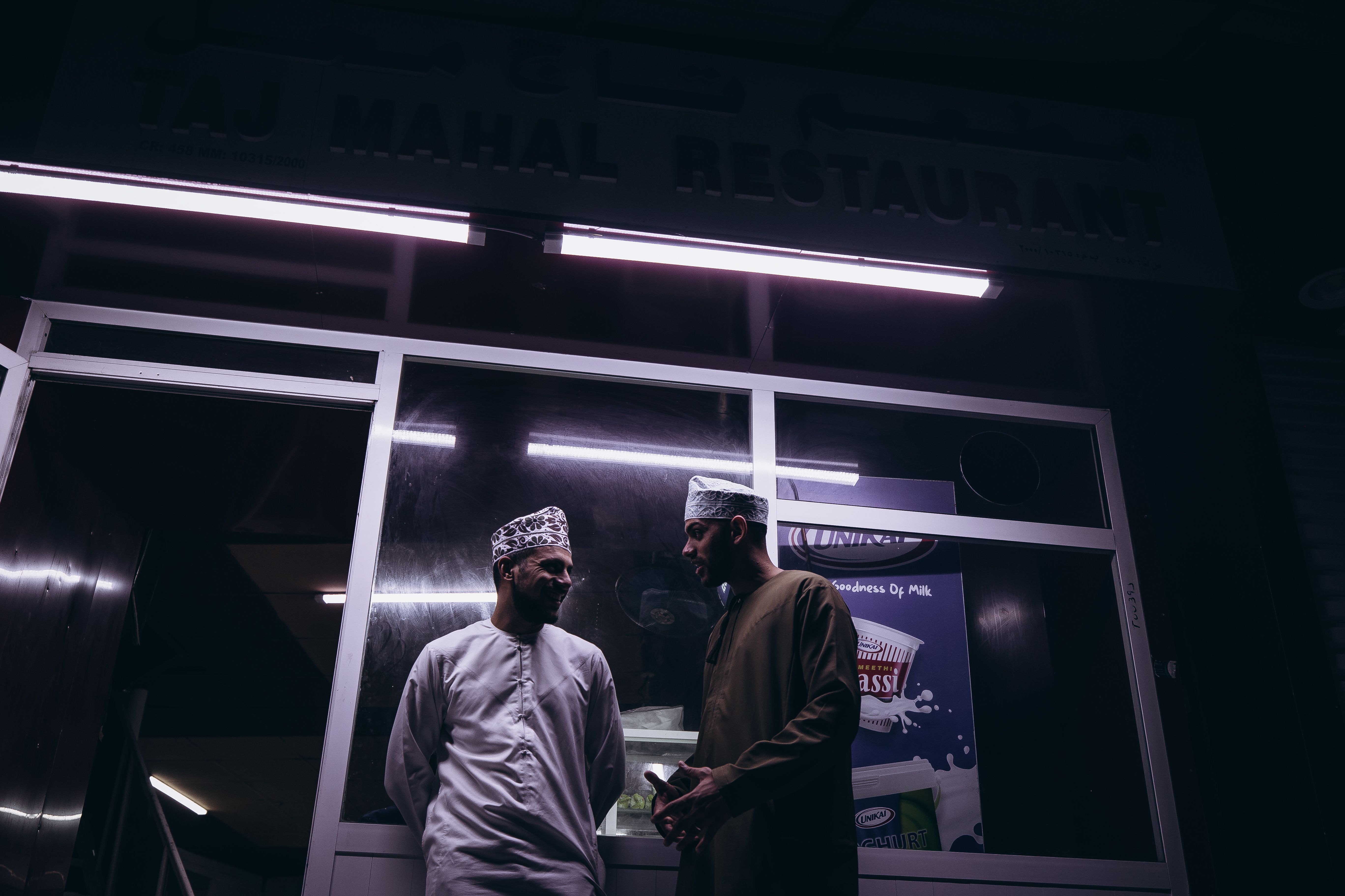 Two men in traditional attire outside a restaurant at night in Muscat, Oman.