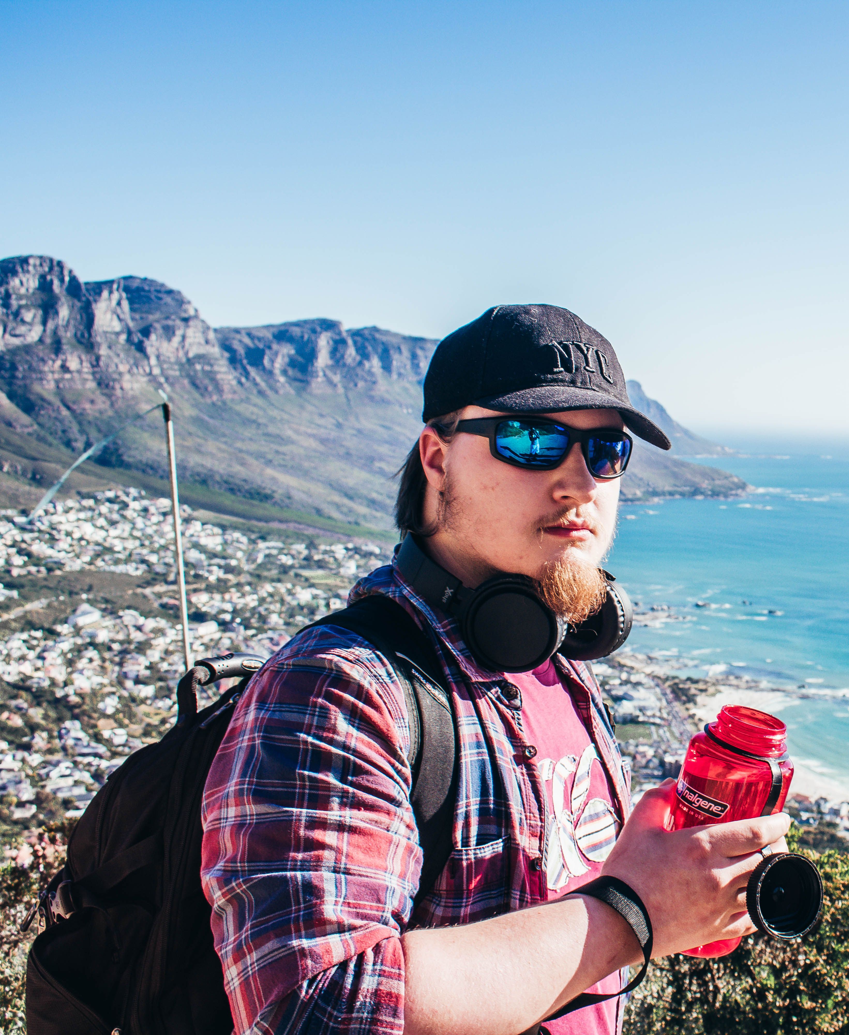 A young man with sunglasses and headphones enjoys a coastal view while holding a red water bottle.