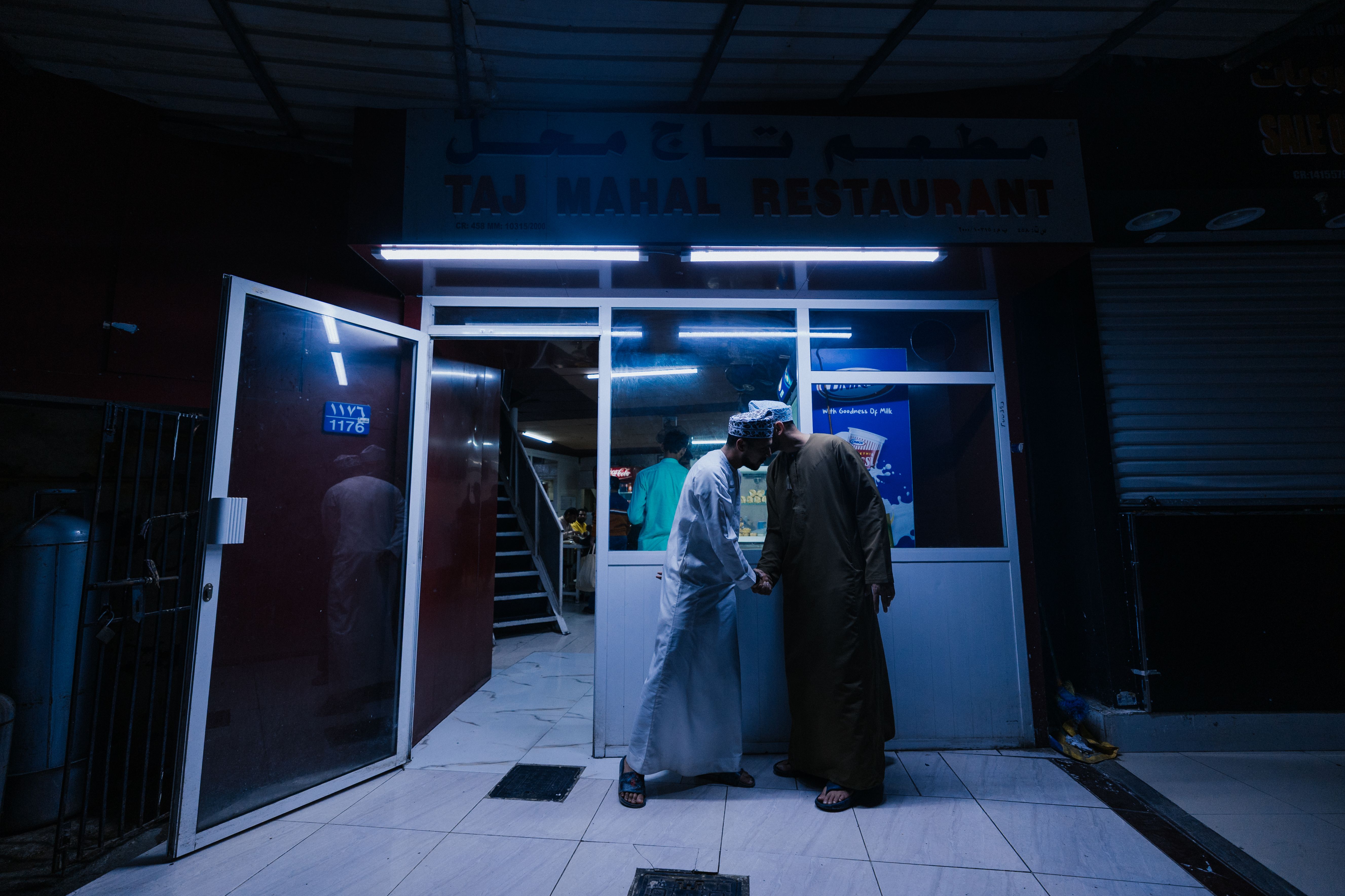 Two men greet each other outside the Taj Mahal Restaurant in Muscat, Oman at night.