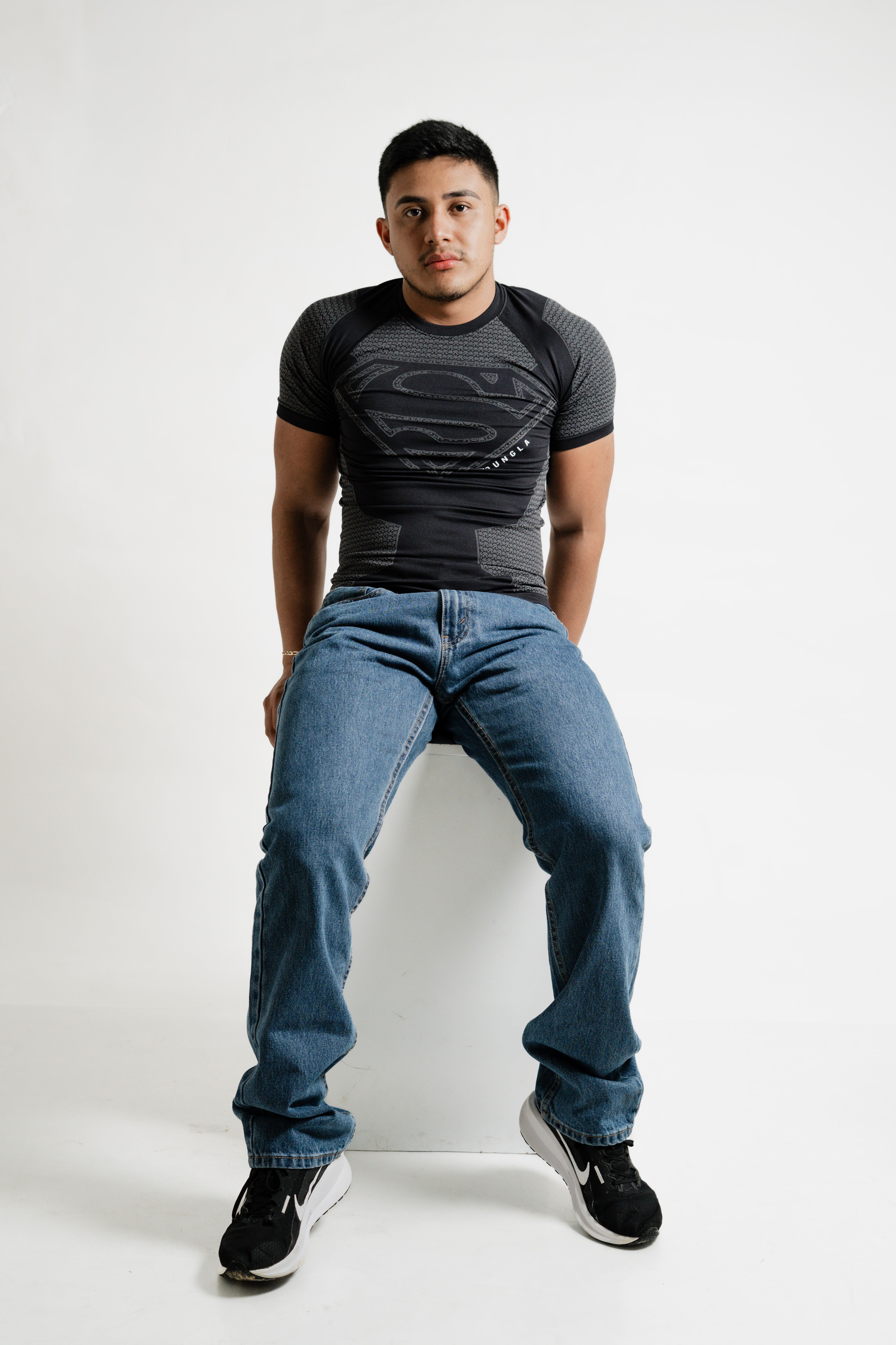 Casual portrait of a young man wearing jeans and a superhero shirt, sitting indoors.