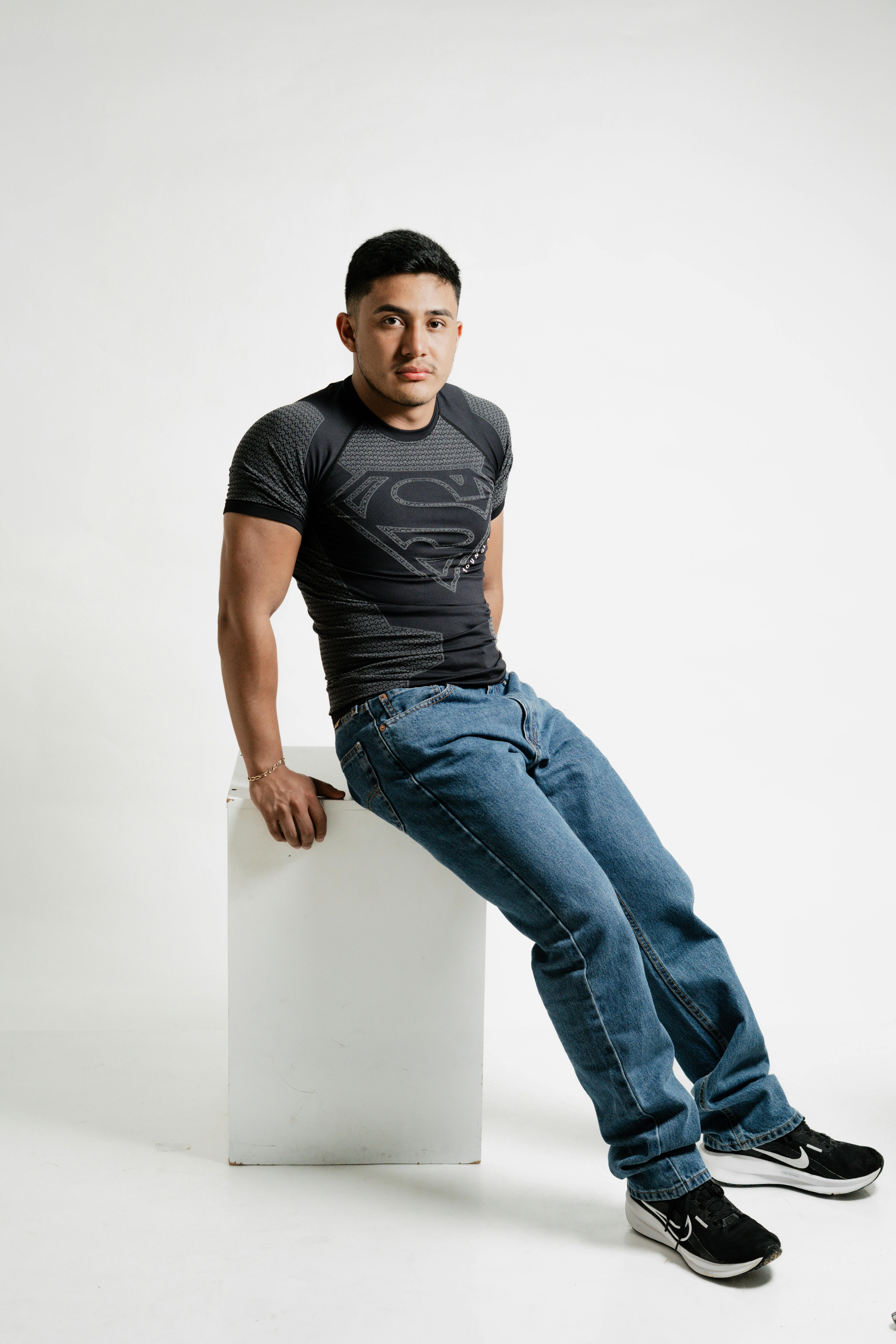Young man poses in a minimalist studio wearing a Superman t-shirt and jeans, exuding casual confidence.