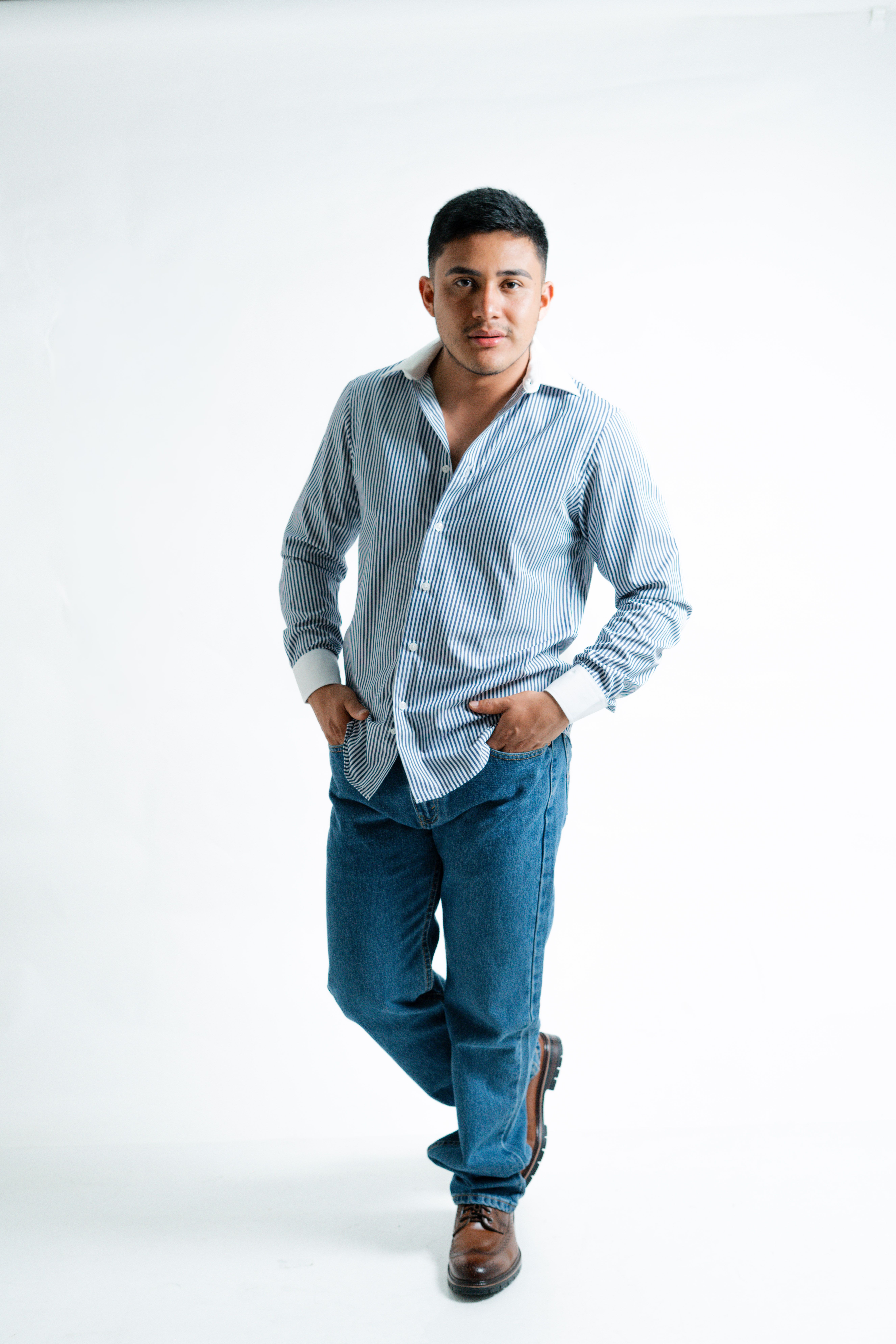 Portrait of a young man in a relaxed pose wearing a striped shirt and jeans in a studio.
