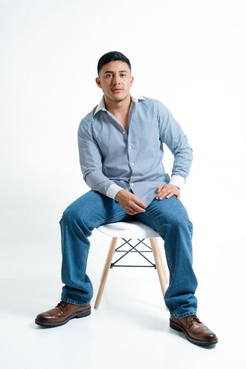 Free Casual portrait of a man sitting on a chair in a studio setting wearing a striped shirt and jeans. Stock Photo