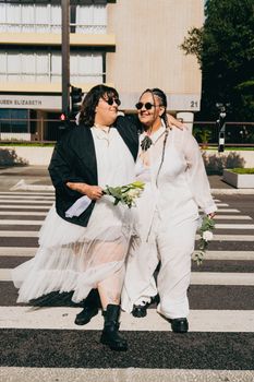 Two adults in joyful embrace, wearing stylish outfits, walking across a city crosswalk on a sunny day.