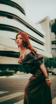 Stylish woman with red hair poses confidently in front of modern architecture.