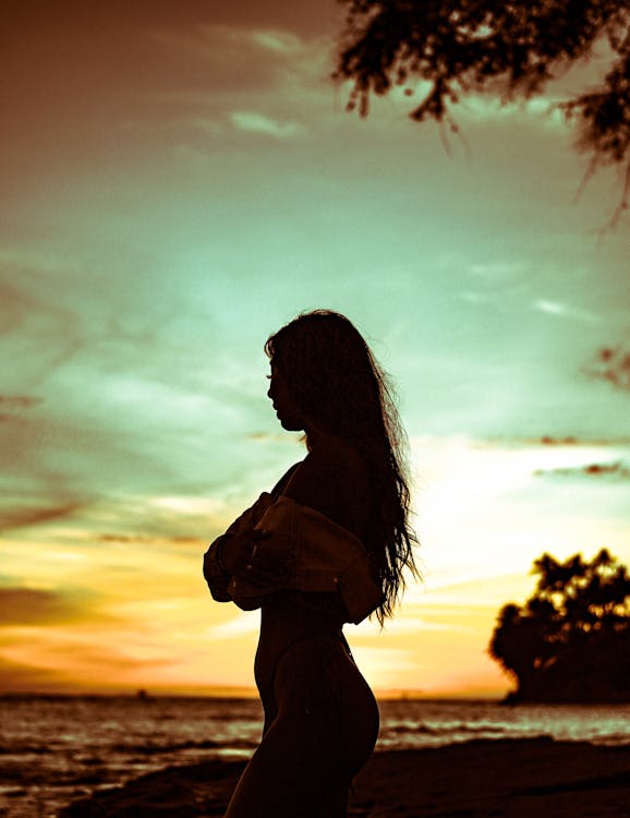 Side view silhouette of anonymous slim woman in swimwear standing on sandy coastline during sunset against cloudy colorful sky