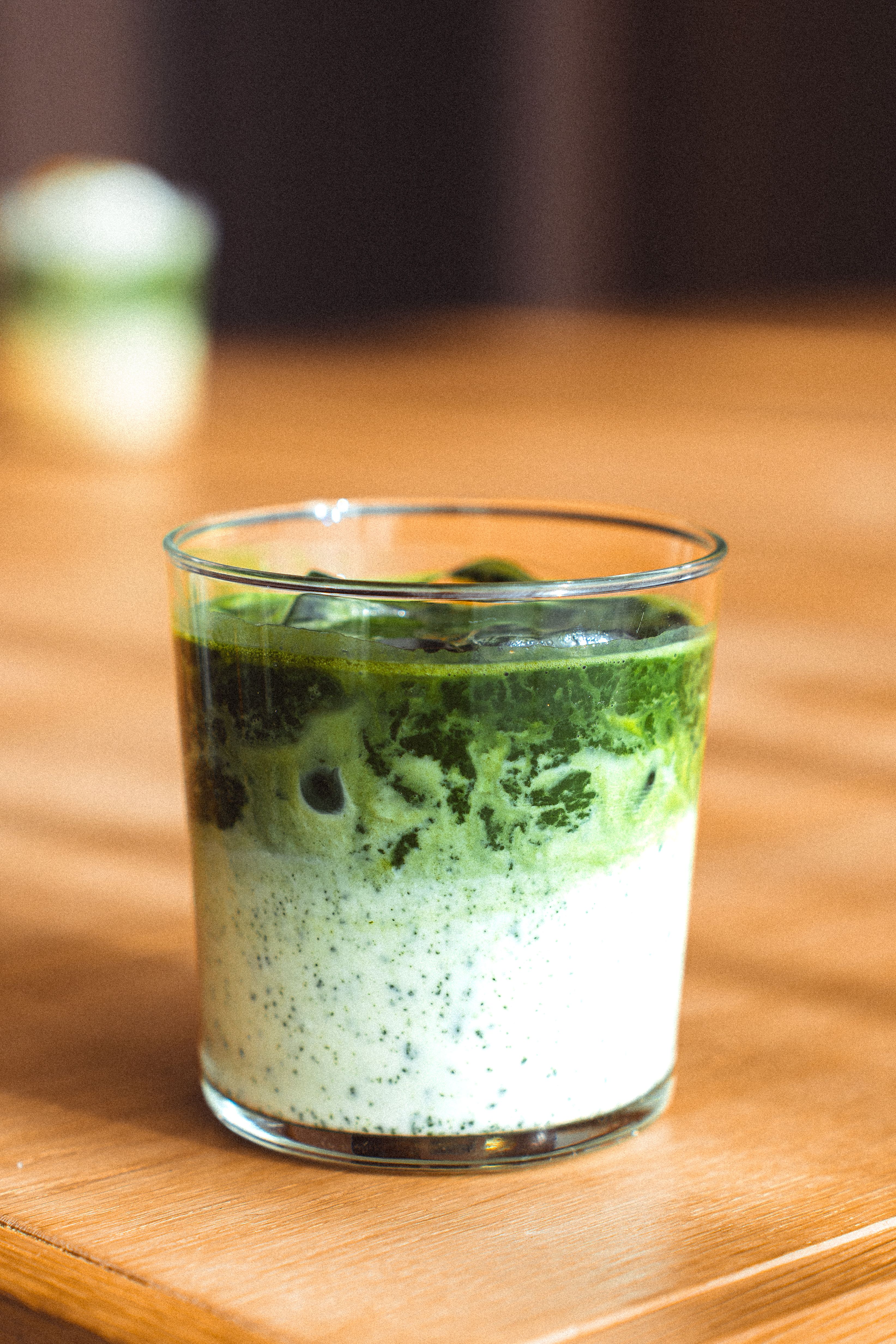 A pale green matcha latte with a thin layer of foam in a clear glass, next to a bamboo whisk and small bowl of matcha powder.