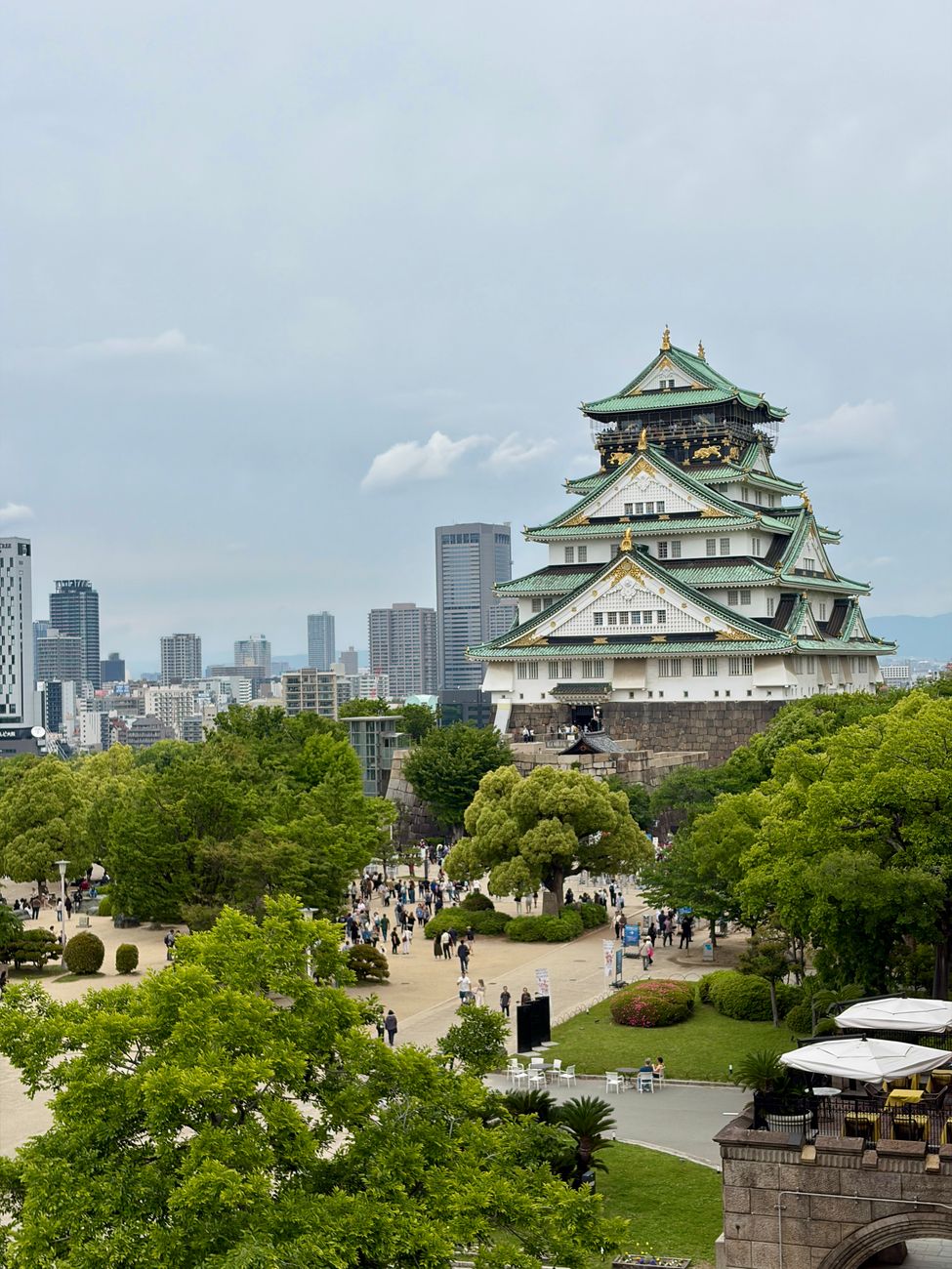 Scenic view of Osaka Castle surrounded by lush greenery and urban skyline on a cloudy spring day.