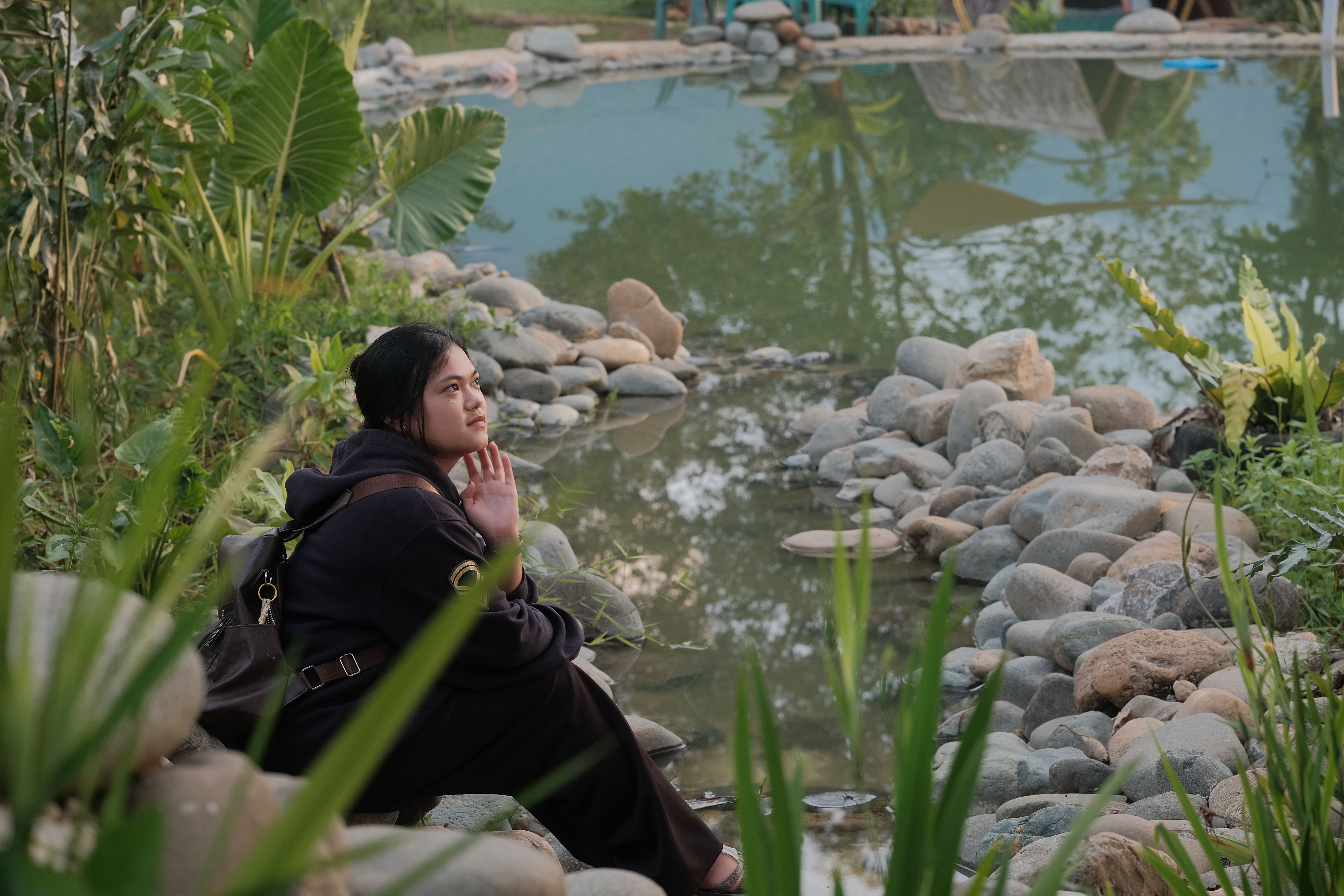 A woman enjoys a peaceful moment by a reflection pond surrounded by lush greenery in Jantho, Aceh.