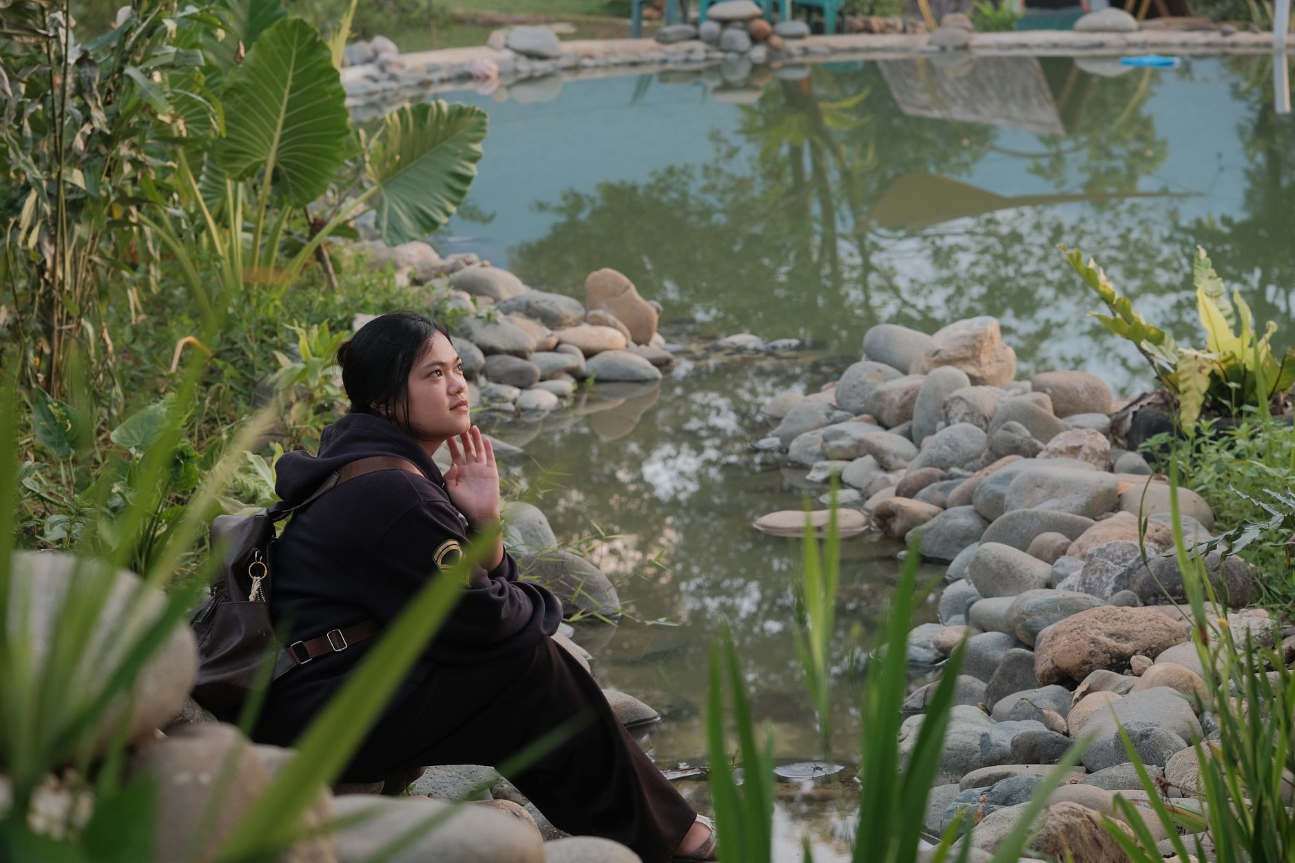 A woman enjoys a peaceful moment by a reflection pond surrounded by lush greenery in Jantho, Aceh.