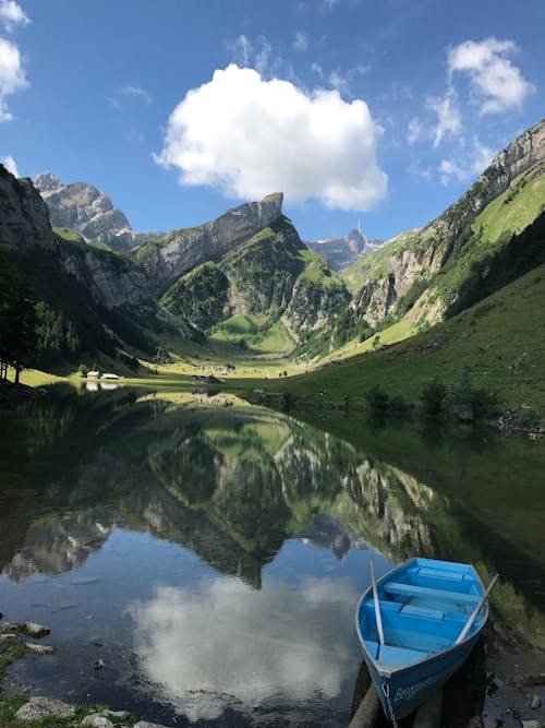 Calm lake with boat on shore surrounded by green mountains under blue cloudy sky