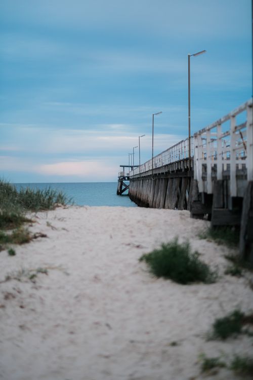 Free Tranquil scene of a sandy beach leading to a rustic wooden pier under a calm evening sky. Stock Photo