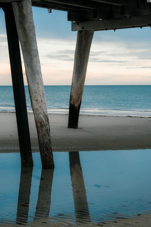 Free Serene view of a calm beach with pier support beams mirrored in wet sand. Perfect for travel and nature themes. Stock Photo