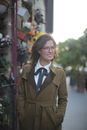 Thoughtful smiling female in jacket and elegant blouse looking away with interest and thinking while standing with hands in pockets flower shop in downtown