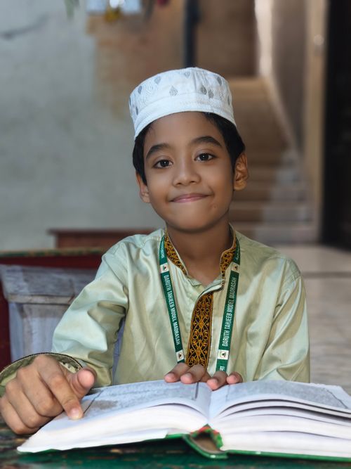 Free Portrait of a young boy in traditional attire reading Quran indoors, conveying education. Stock Photo