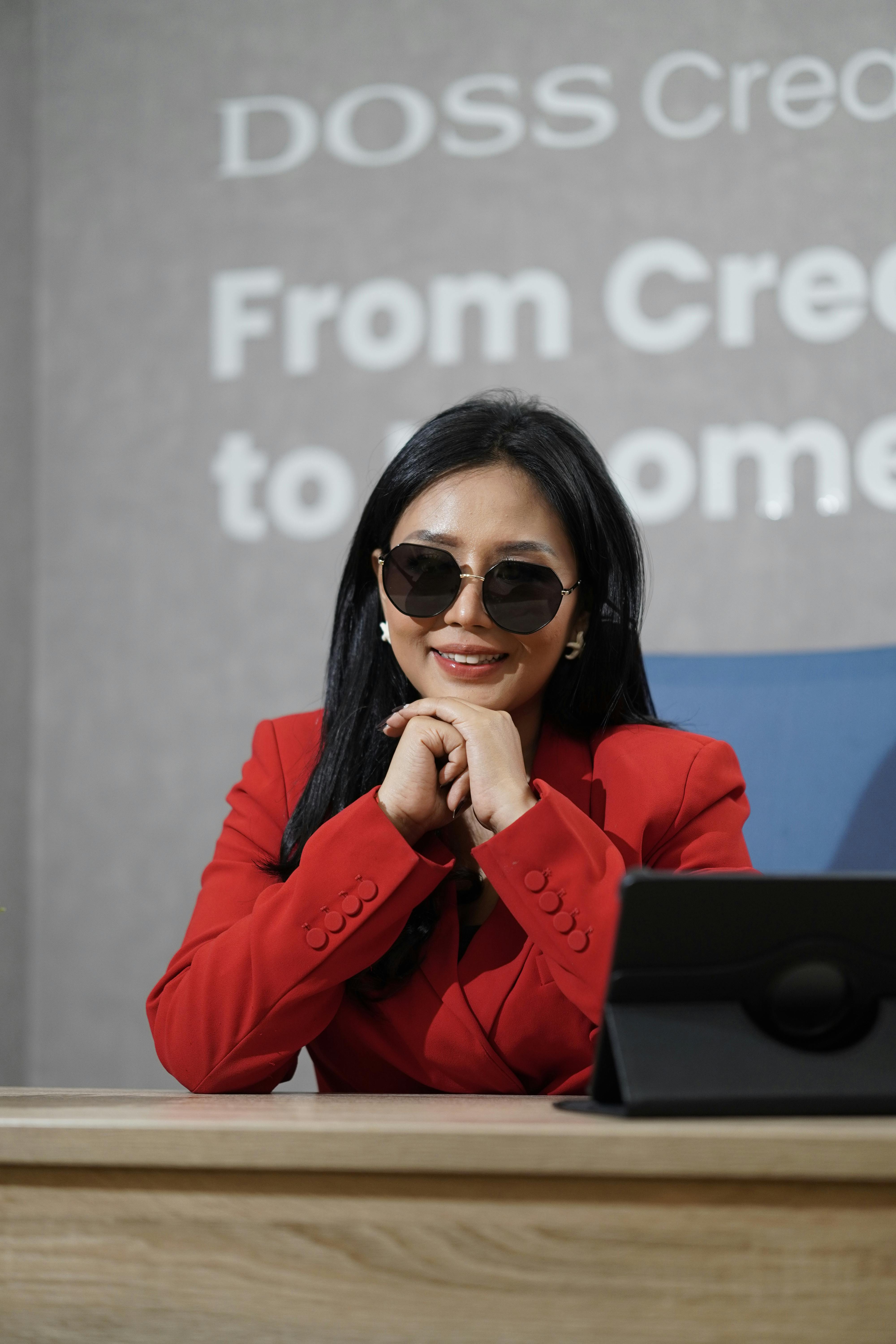 Stylish woman with sunglasses in red blazer behind desk, indoors.