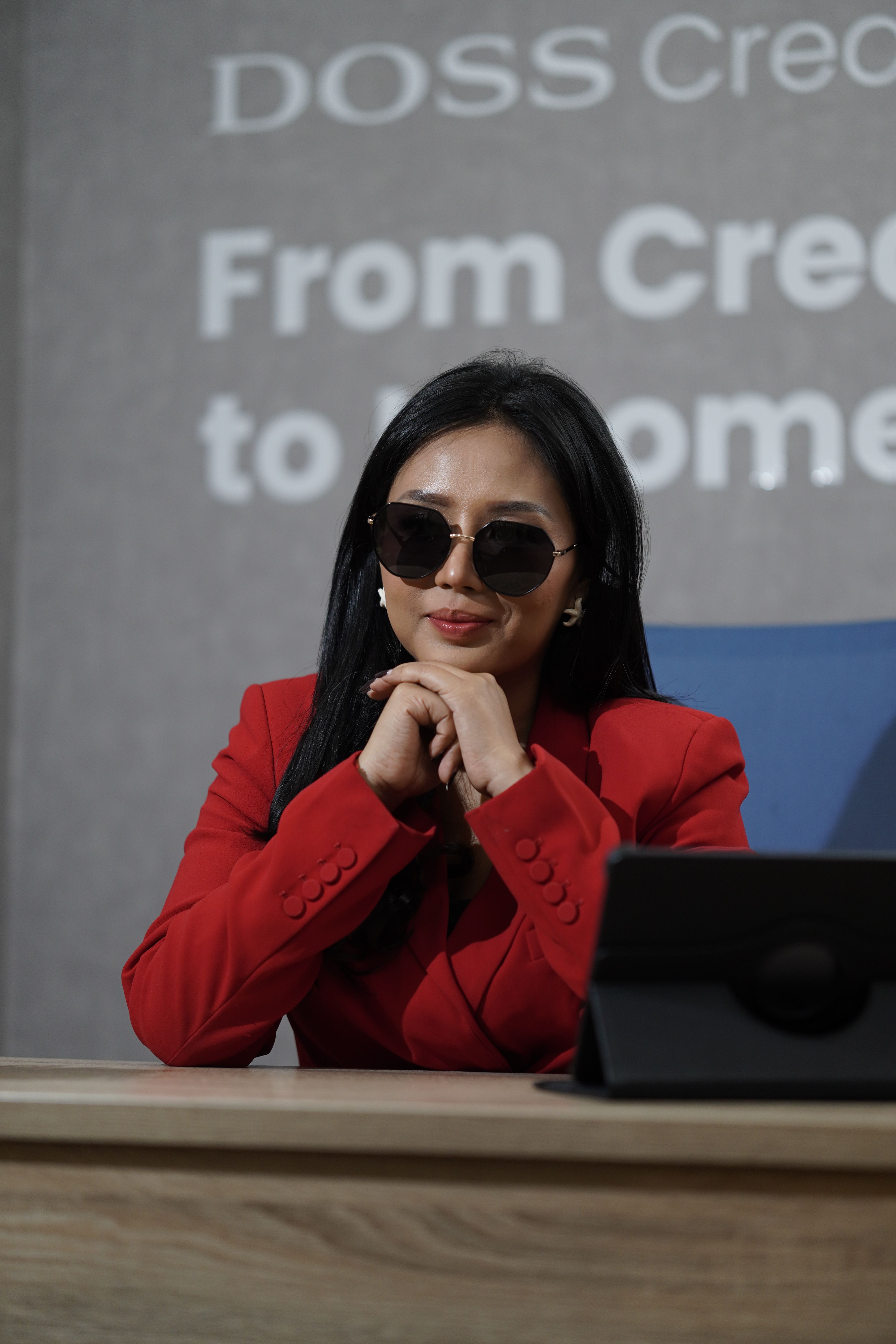 Businesswoman wearing a red blazer and sunglasses sits confidently at a desk indoors.