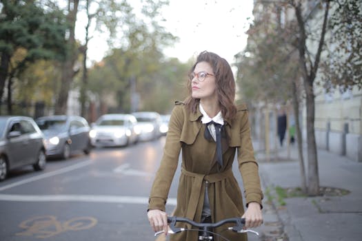 Woman in brown coat bicycling through a city street with cars in background.
