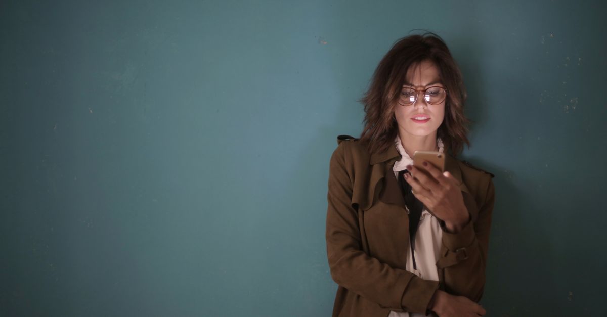 A woman with eyeglasses and brown hair uses a smartphone against a teal wall indoors.
