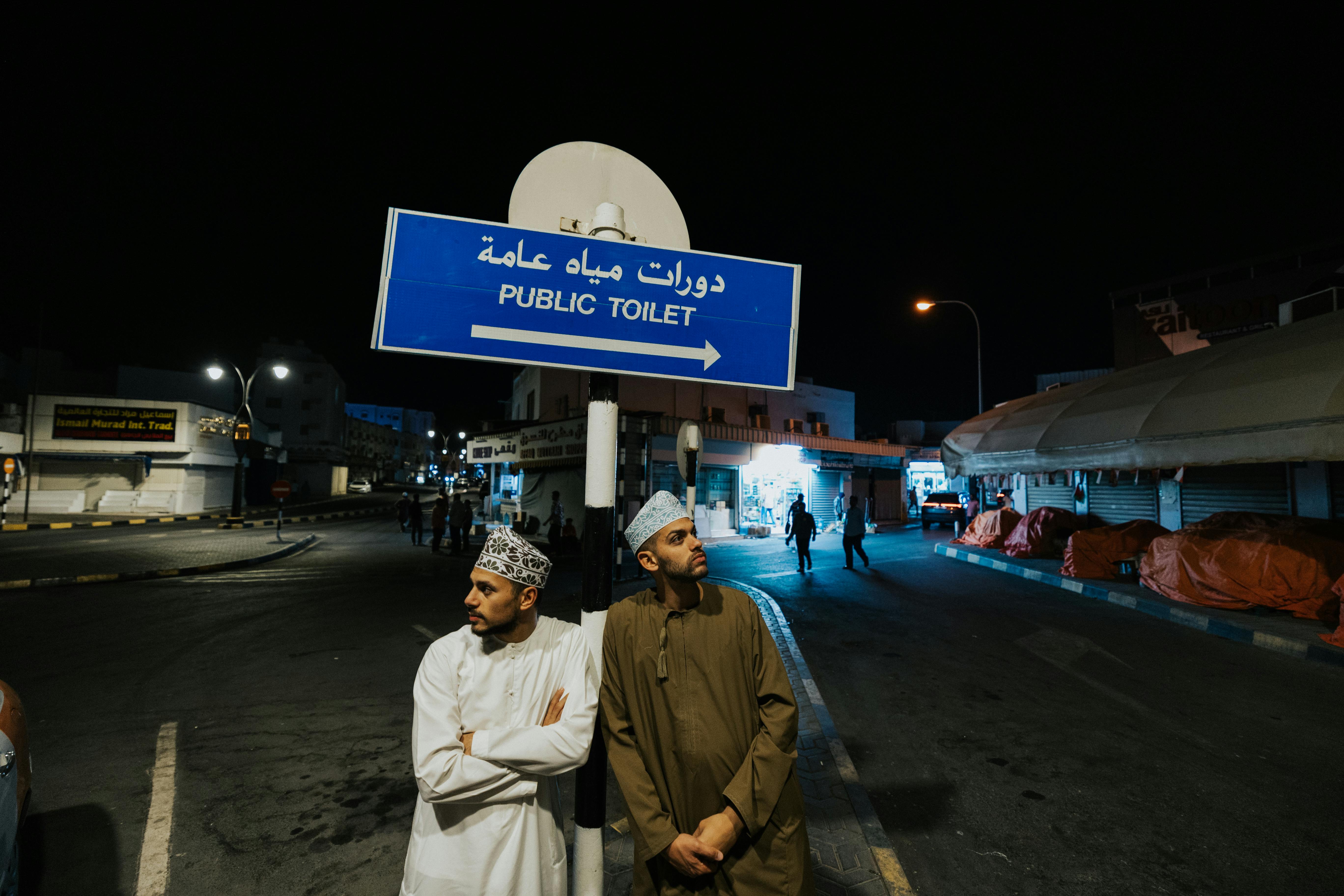 Two men in traditional attire stand under a public toilet sign in Muscat, Oman at night.