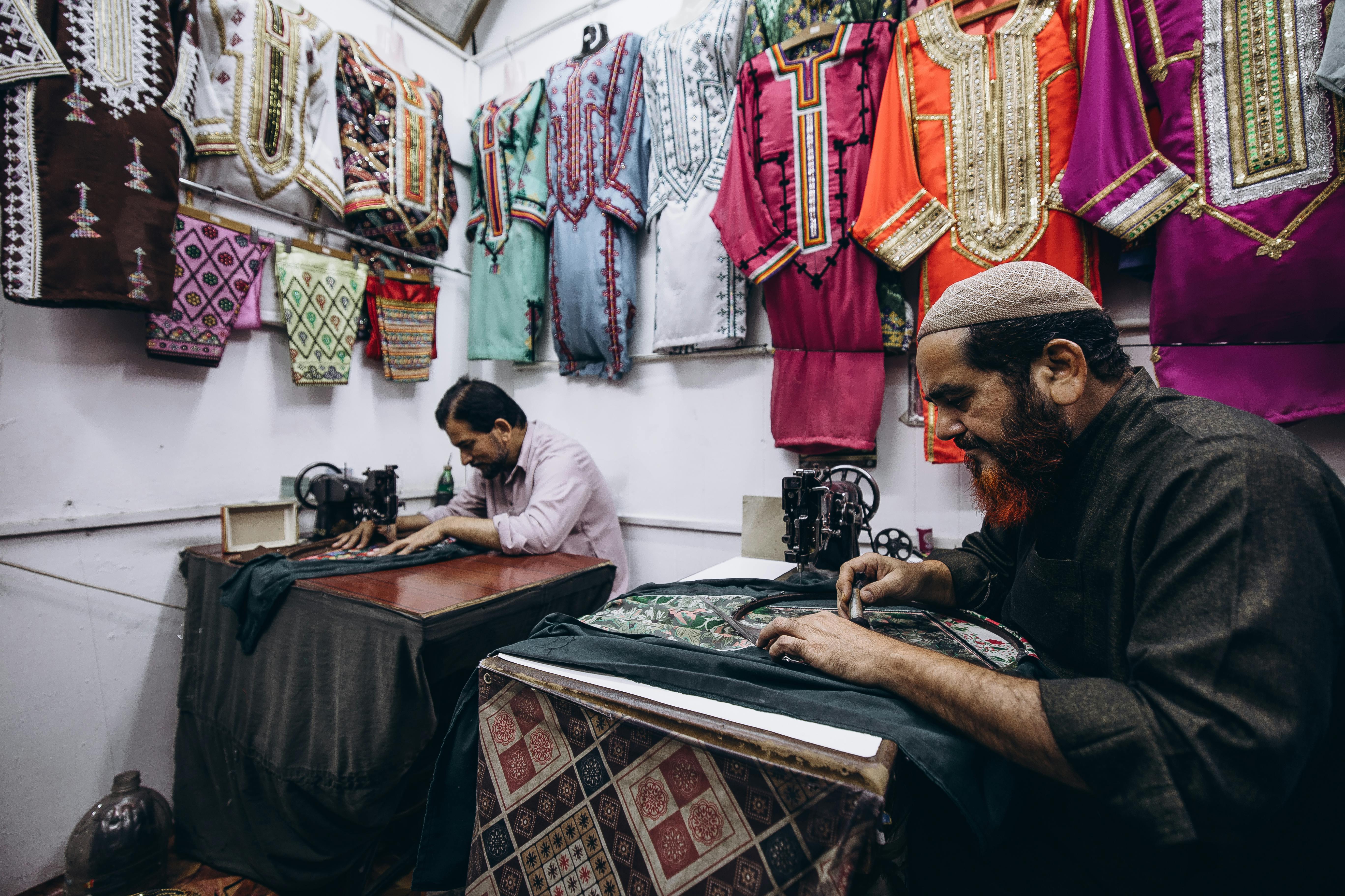 Two tailors crafting garments with traditional attire displayed in a Muscat shop