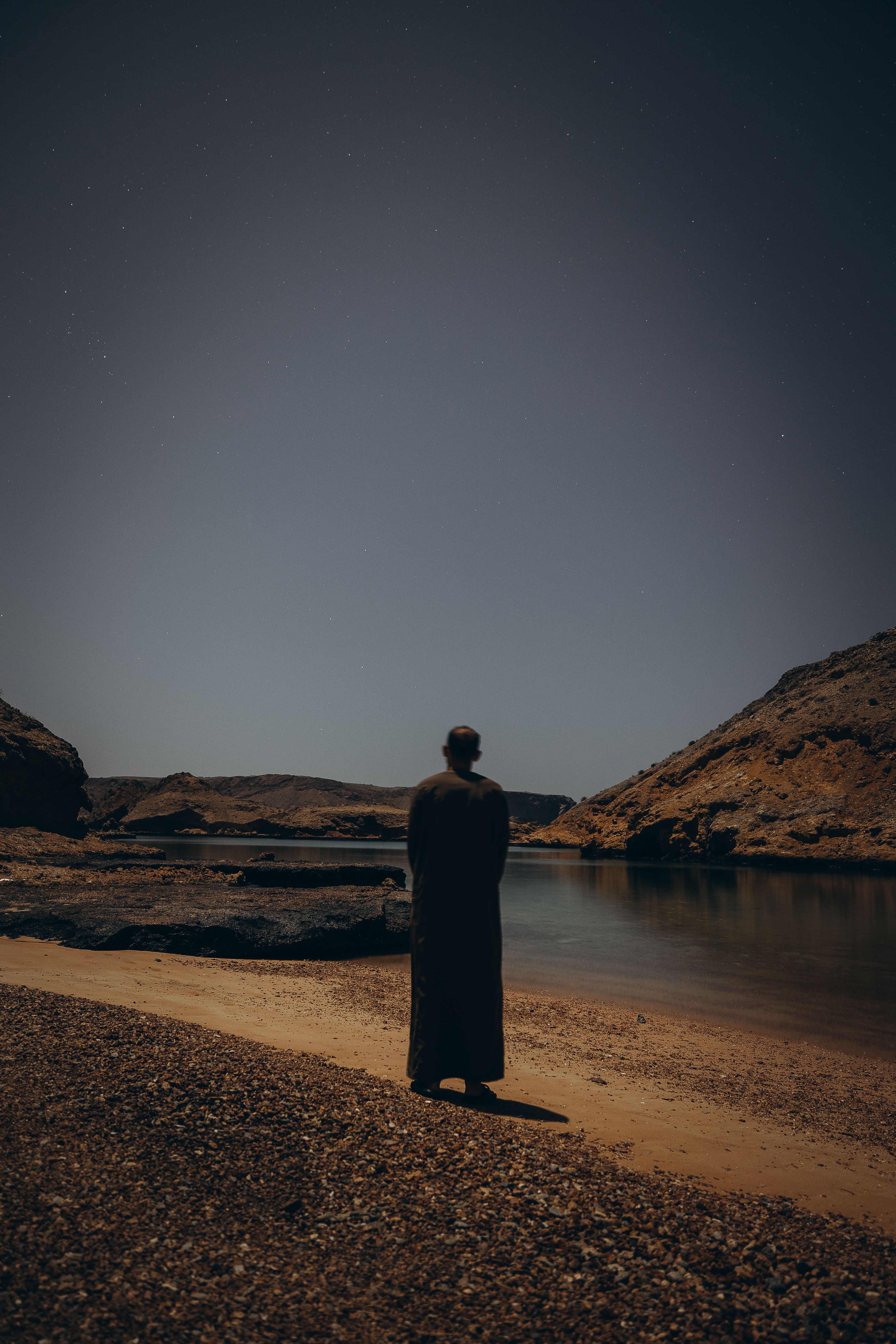 A lone figure stands on a tranquil beach in Muscat, Oman, under a starry sky during twilight.