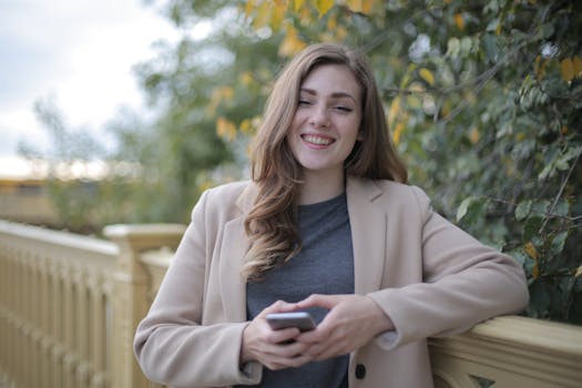 A cheerful woman in a blazer smiles while holding a smartphone outdoors.