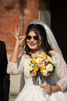 Smiling bride poses with sunglasses and colorful bouquet against a rustic backdrop.