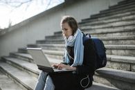 一个女人戴着耳机、背着背包，在户外楼梯上用笔记本电脑在线学习