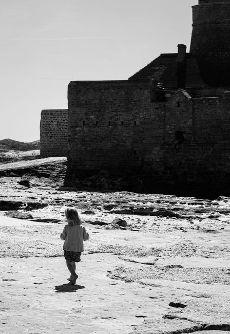A child walks near an old fortress overlooking a rocky shore on a sunny day.