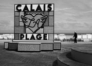 Black and white photo of Calais La Plage sign with cloudy skies and a lone person walking.
