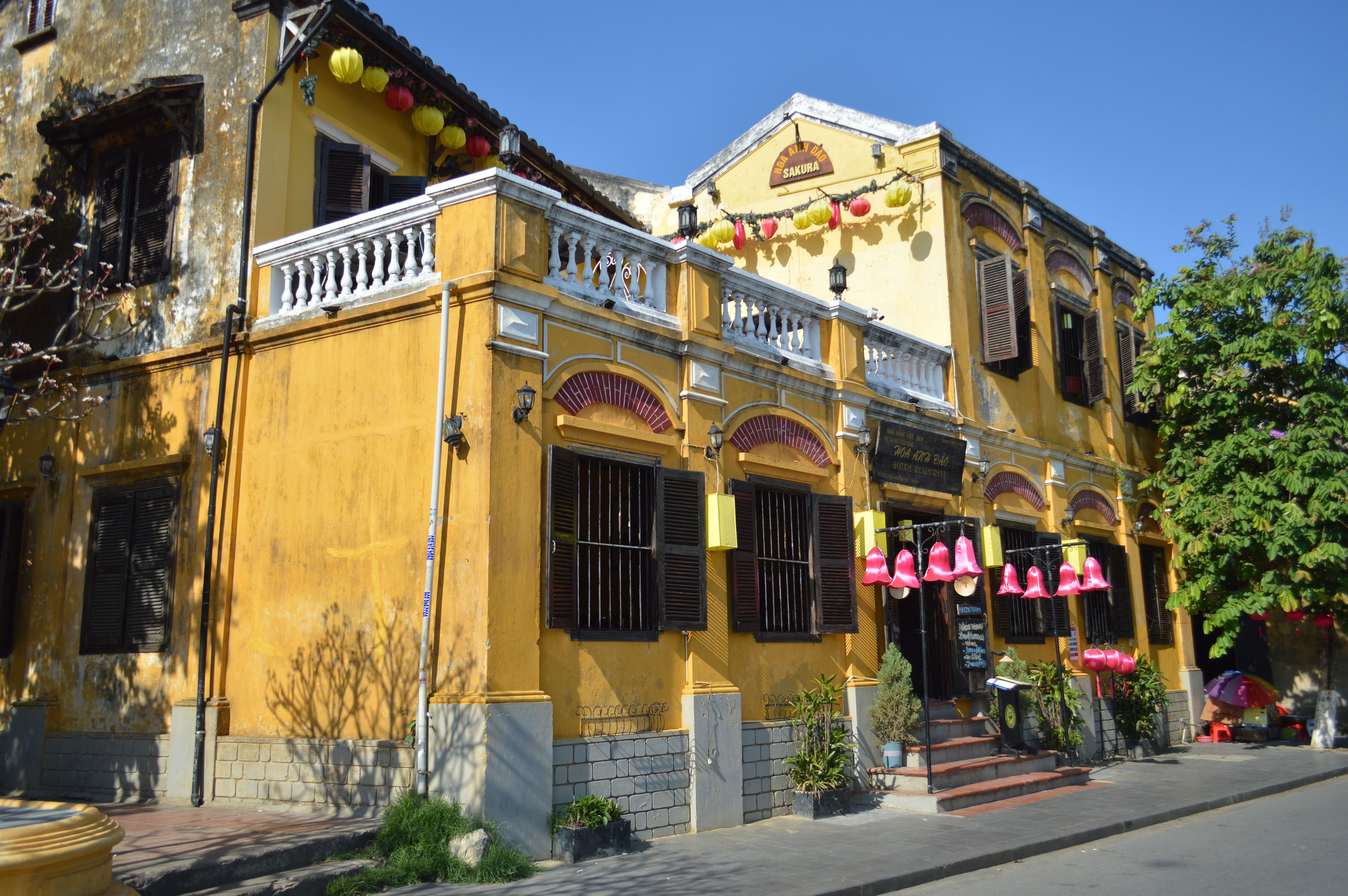 A yellow two-storey colonial-style building with a balcony and shuttered windows, with strings of pink lanterns outside and trees beside it