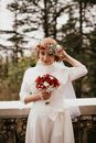 一位头戴花冠、身着白色婚纱的新娘，在户外手持红白相间的花束，脸上带着微笑