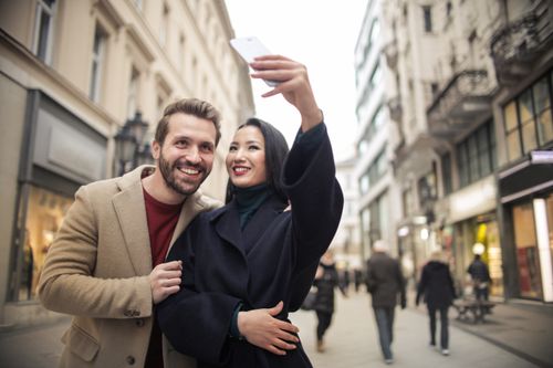 Free Happy couple takes a selfie on a lively urban street in daylight, capturing joyful moments during their city walk. Stock Photo