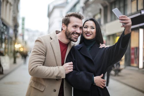 Man in Brown Coat Smiling Beside Woman in Black Coat Taking Selfie