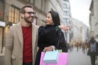 一位快乐时尚的亚洲女人穿着温暖的衣服，微笑着与一位开朗的未刮胡子的戴眼镜男人，在购物后一起走在城市街道上，手指移开