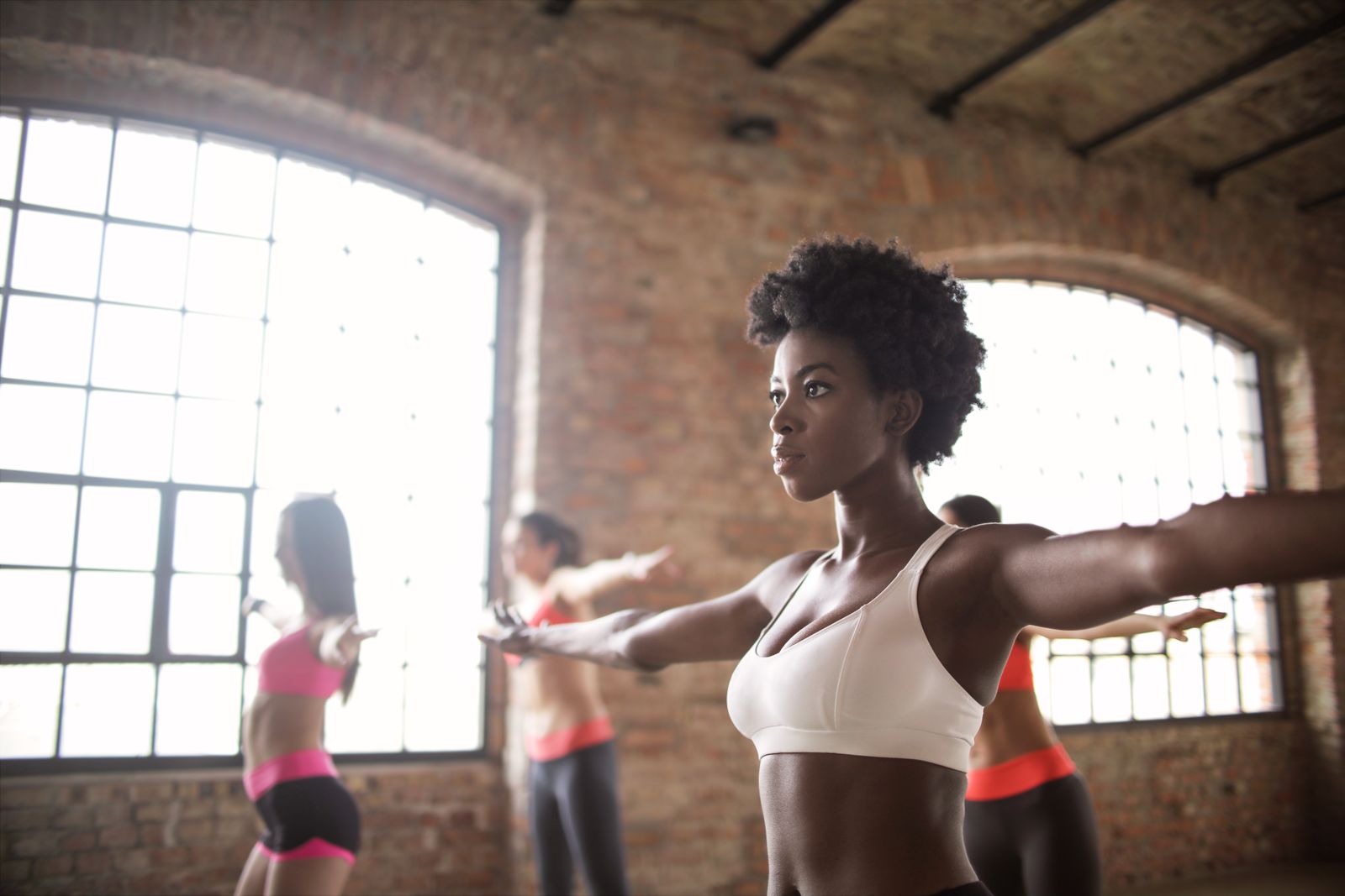 Woman in white sports top stretches arms wide in a sunlit brick studio with large windows