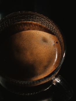 Dark moody close-up of frothy espresso in a clear glass cup creates a cozy ambiance.
