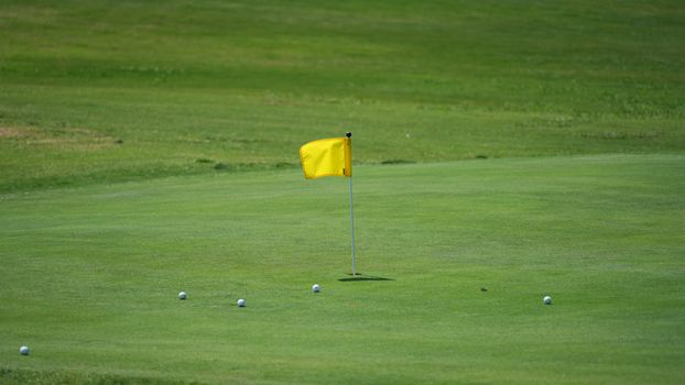 Golf course scene with yellow flag and golf balls on green under a clear sky.