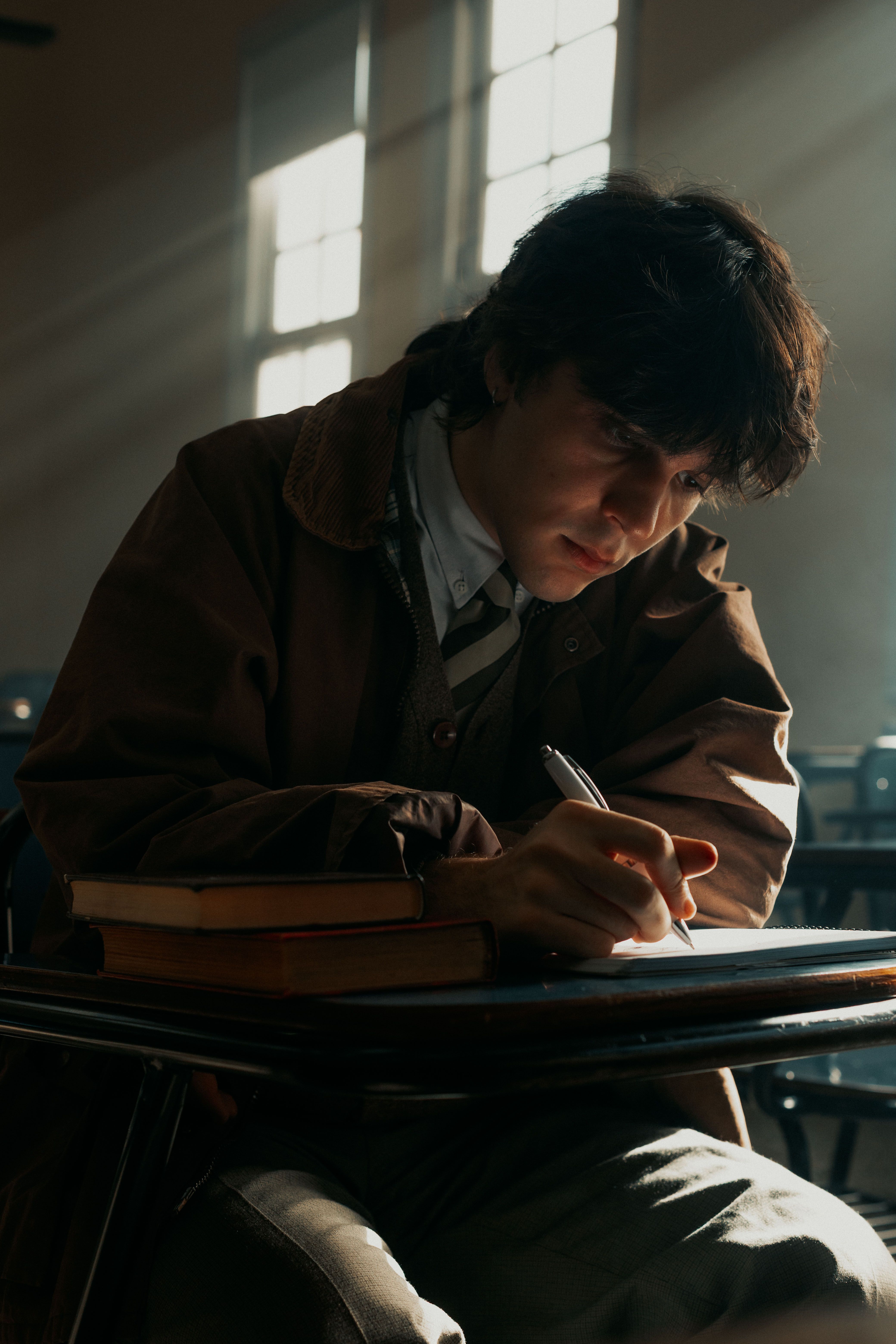 A student intense in thought, writing with focus in a dimly lit classroom with natural light streaming in.