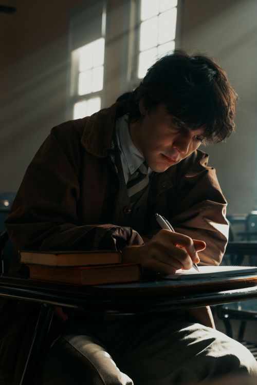 Free A student intense in thought, writing with focus in a dimly lit classroom with natural light streaming in. Stock Photo