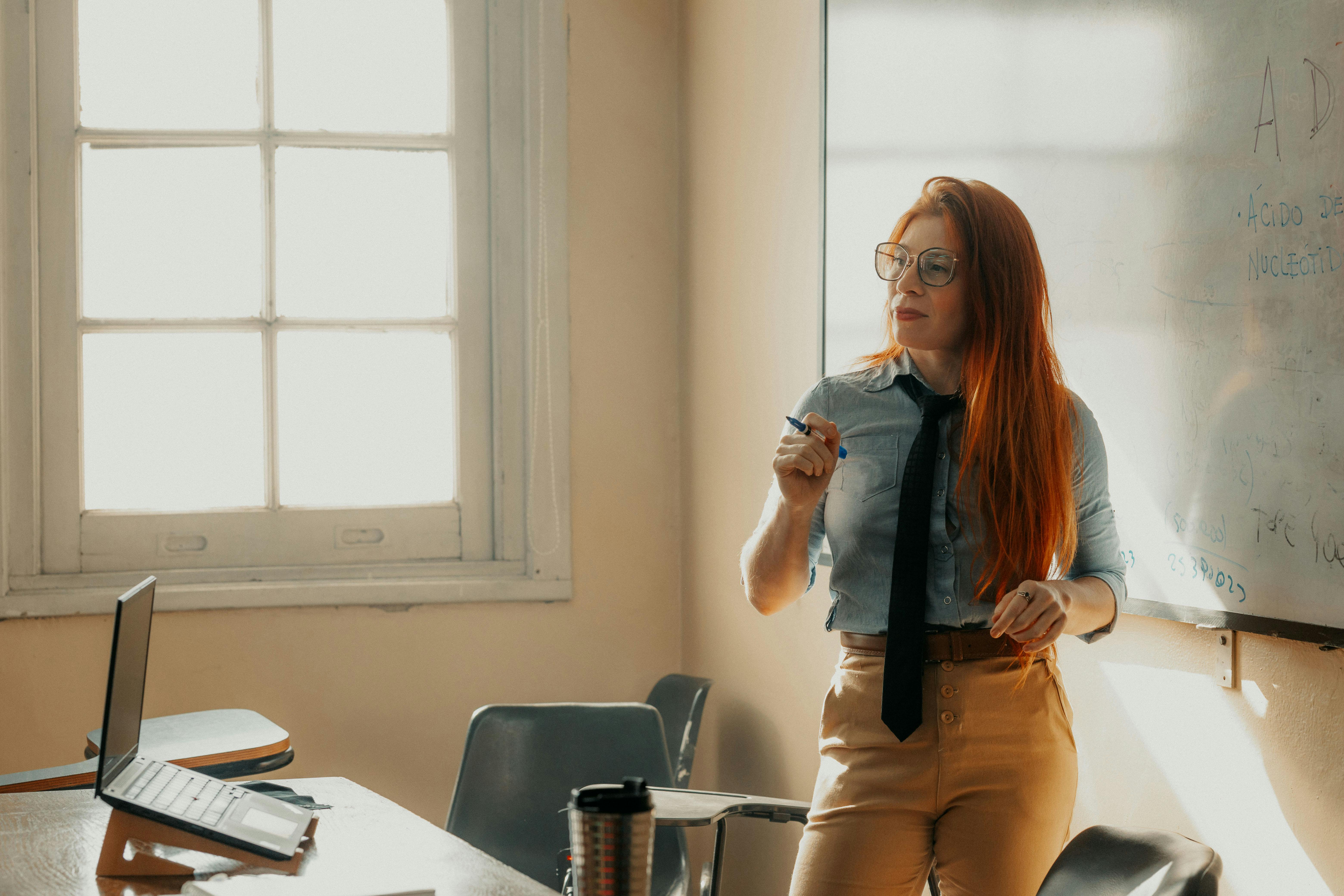 A female lecturer teaches in a sunny classroom, pointing at a whiteboard with notes.