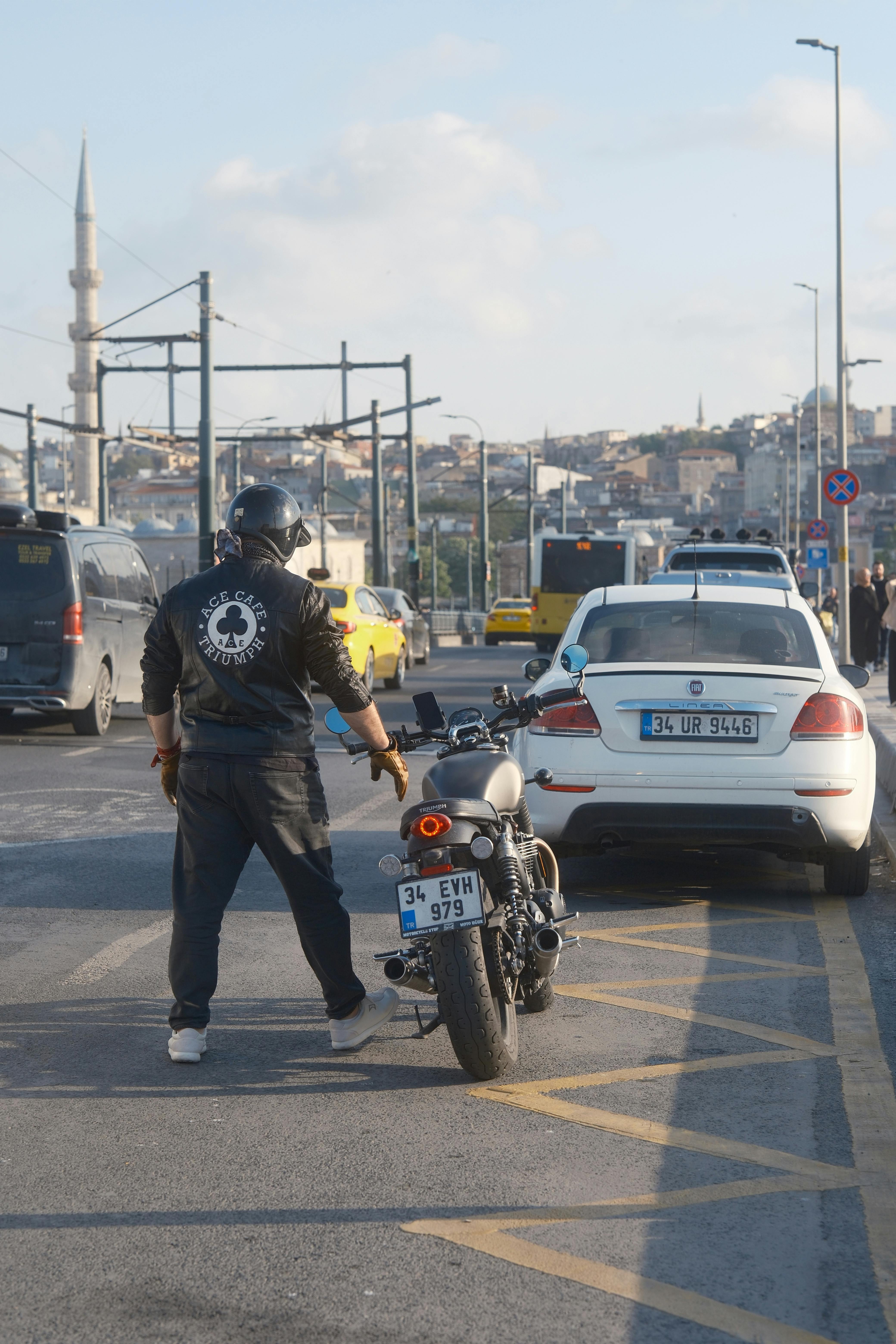 A motorcyclist stands near a parked motorcycle on a busy street with cars and cityscape in the background.